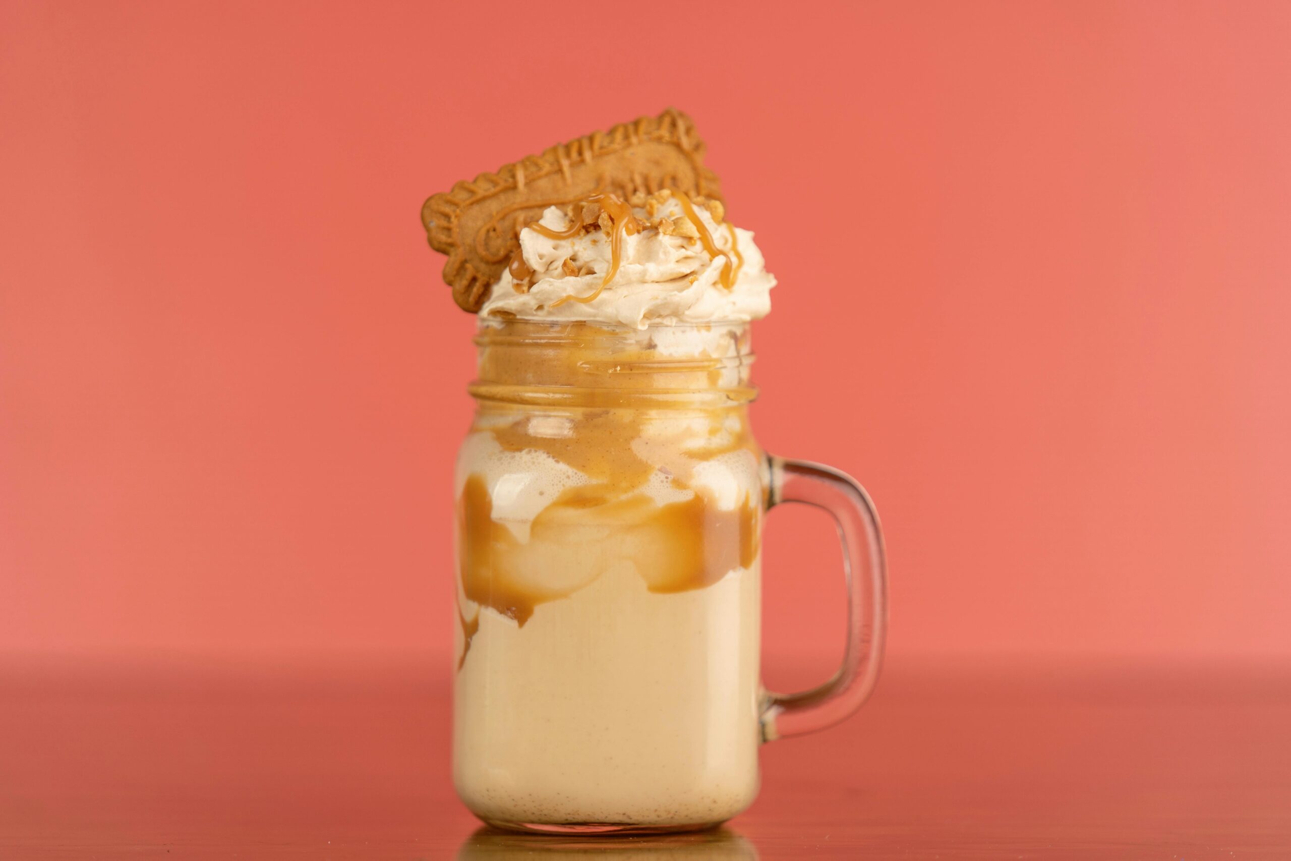 Close-up of a caramel milkshake with whipped cream and biscuit topping against a salmon background.